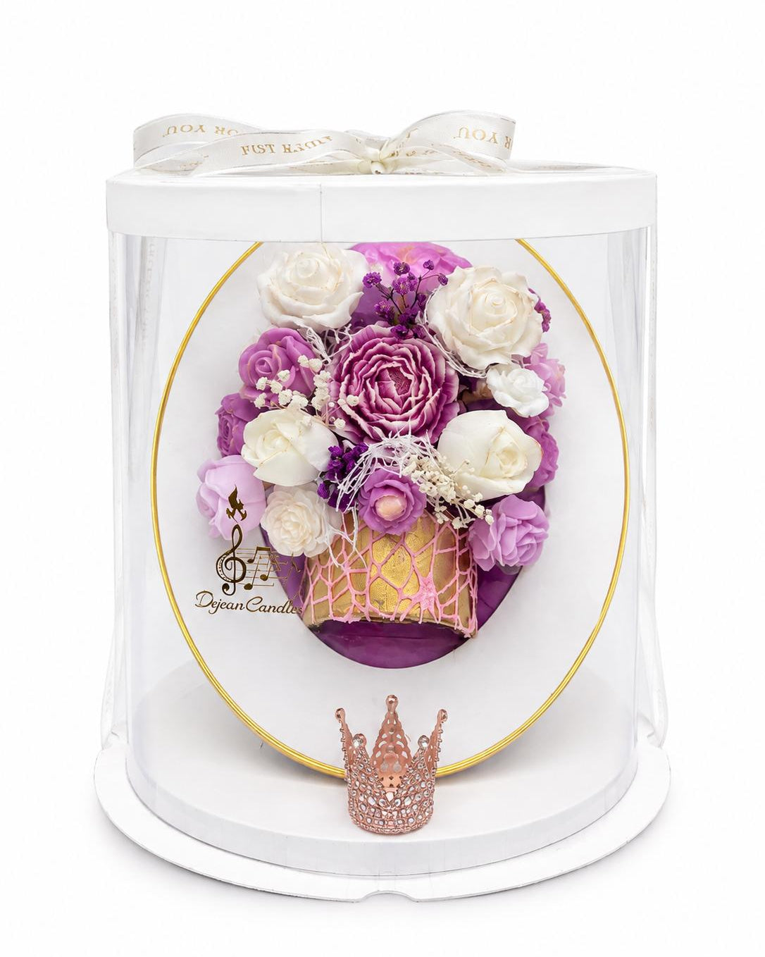 A preserved flower arrangement in a clear cylindrical gift box with a white lid and ribbon. The bouquet features purple and white roses, a gold crown, and a small pink glitter crown in front.